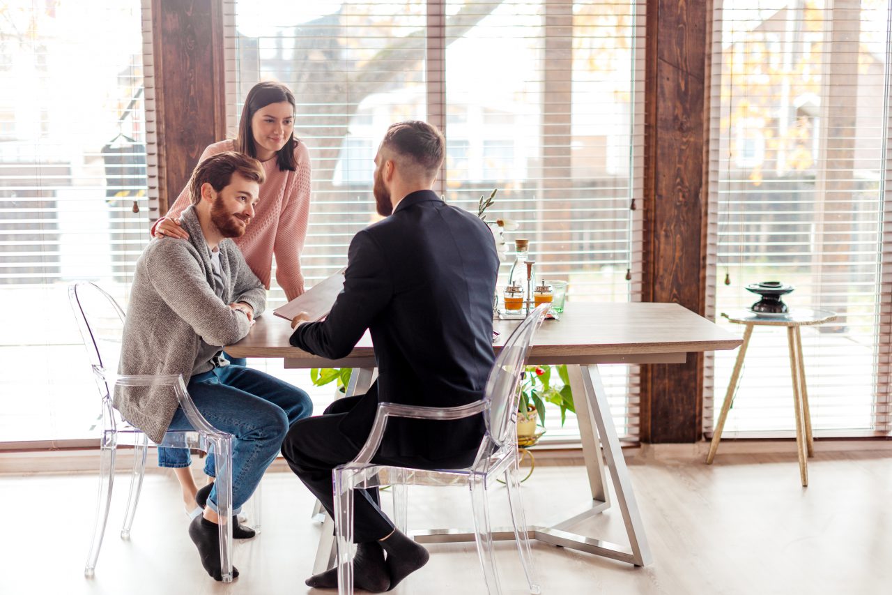 Young caucasian couple consulting with bank financial adviser before buying new house. Two bearded men sitting at table and checking documents while woman stands behind.